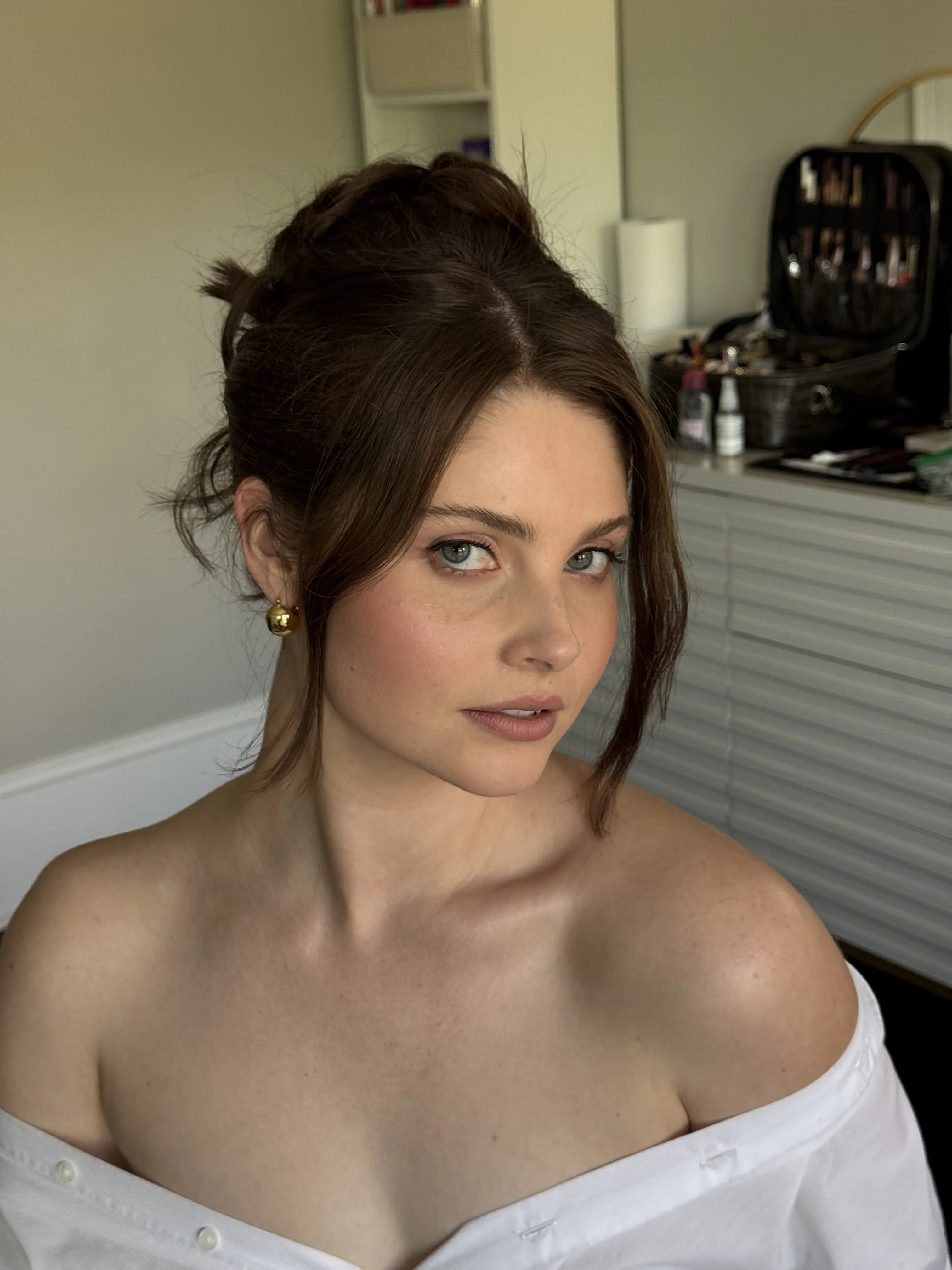 A bride with a soft updo during hair and makeup, off the shoulder white shirt